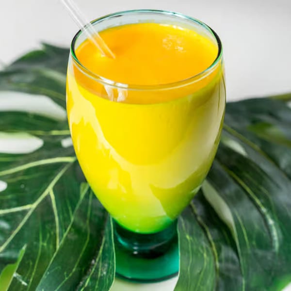 Bright yellow mango lassi in a green-stemmed glass with a straw, on large green leaves.