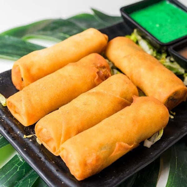 Crispy golden spring rolls on a black plate with green and brown dipping sauces, garnished with green leaves.